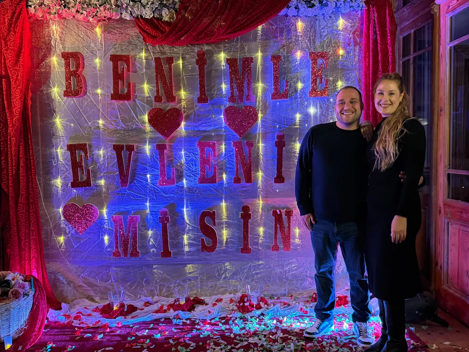 A couple in front of a lit sign and red curtains after a marriage proposal in Edirne